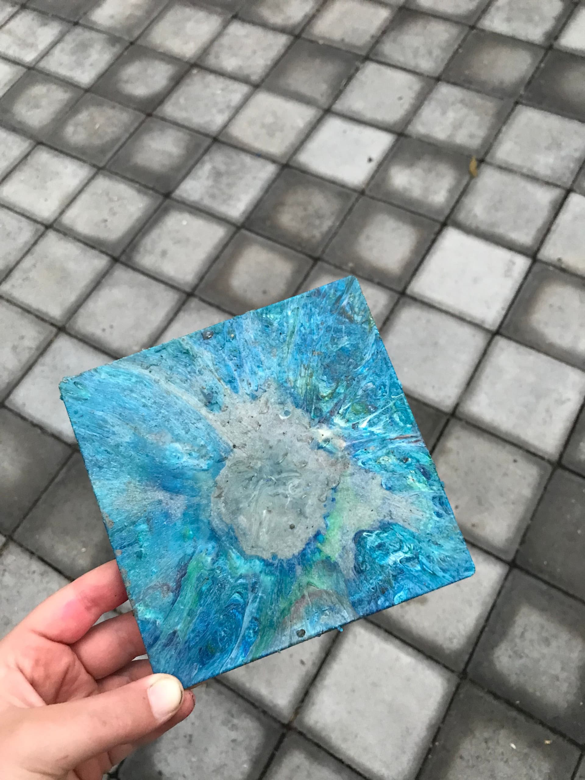 A hand holding a square tile with a blue and green abstract design made from recycled material against a background of gray paving stones.